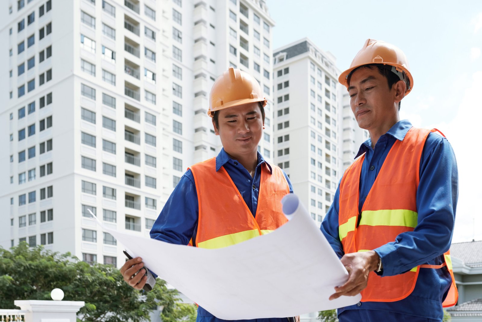 Smiling Asian construction workers meeting outside building and discussing blueprint